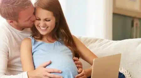 זוג בהריון יושבים ביחד על הספה עם לפטופ ומחייכים