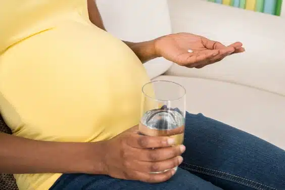 אישה בהריון יושבת על הספה ומחזיקה כוס מים ביד אחת וכדור לבליעה ביד שנייה