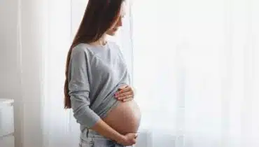 אישה בהריון עם הידיים על הבטן מסתכלת מהחלון